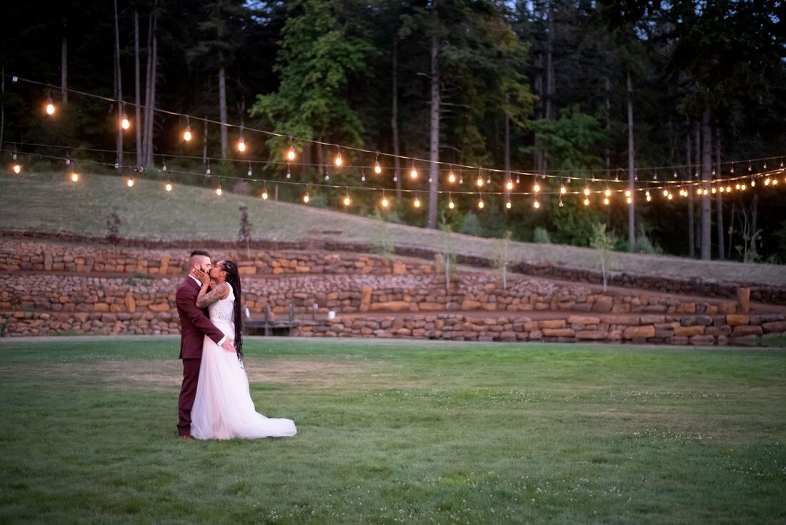 Bride and Groom in a yard with a rock wall behind them and lights hanging above them.