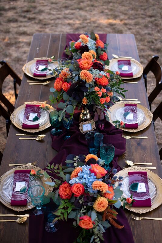 Table with 6 place settings, burgundy table runner, formal place settings with gold charger, bright orange and blue flowers with greenery as centerpieces.