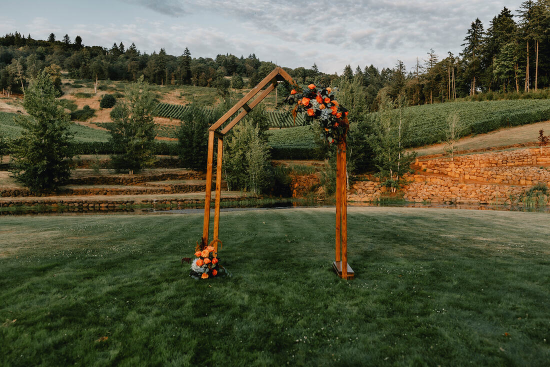 Wedding arch with a vineyrd hill in the background. Arch has orange and blue flowers with greenery on the top.