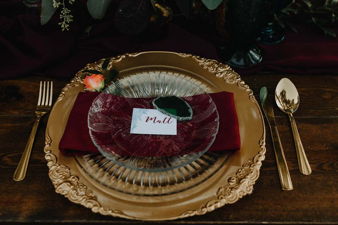 Place setting with clear plate, burgundy napkin, gold charger, gold silverware and place card held in place with green agate.